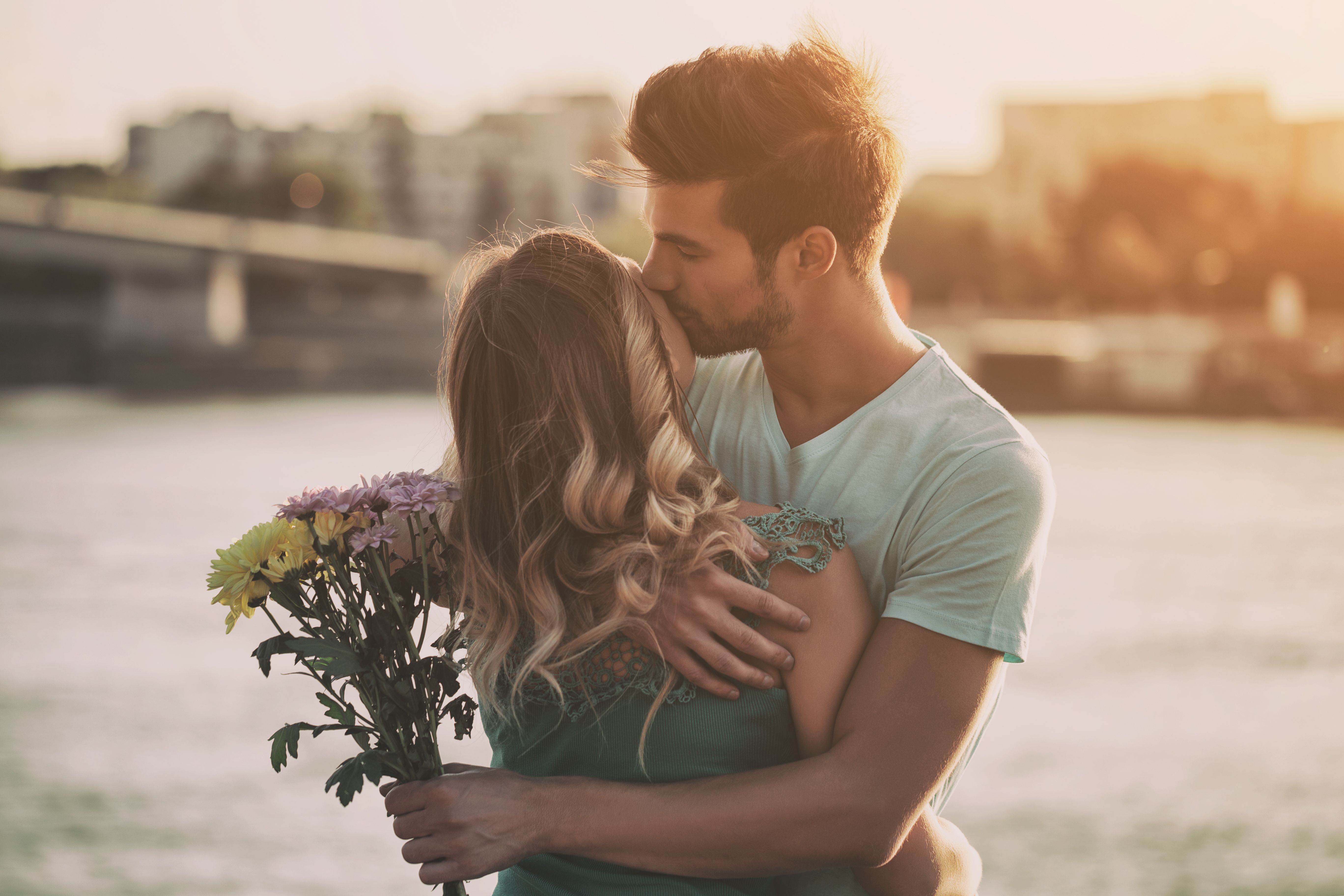 Jeune couple souriant enlacé en ville, l’homme tenant un bouquet de fleurs, illustrant une rencontre sérieuse, une relation durable et un premier rendez-vous réussi.